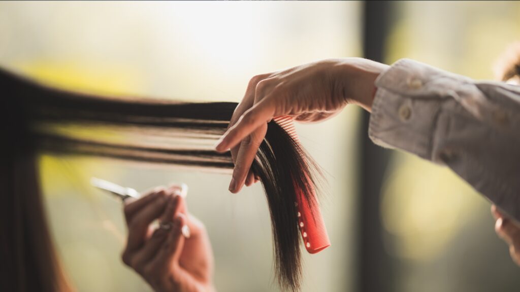 Close-up of hands, akin to those of skilled hairdressers, deftly cutting long, straight hair with scissors and a red comb.