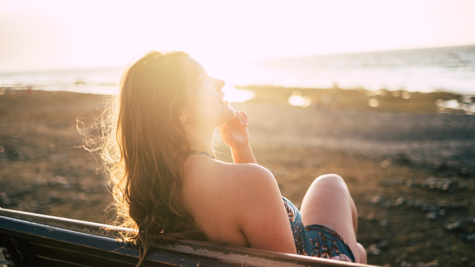 An individual with beautifully coloured hair sits on a beach bench, gazing towards the sun. The sea extends to the horizon, forming a serene backdrop that mirrors their vibrant hairstyle, expertly crafted by skilled hairdressers.